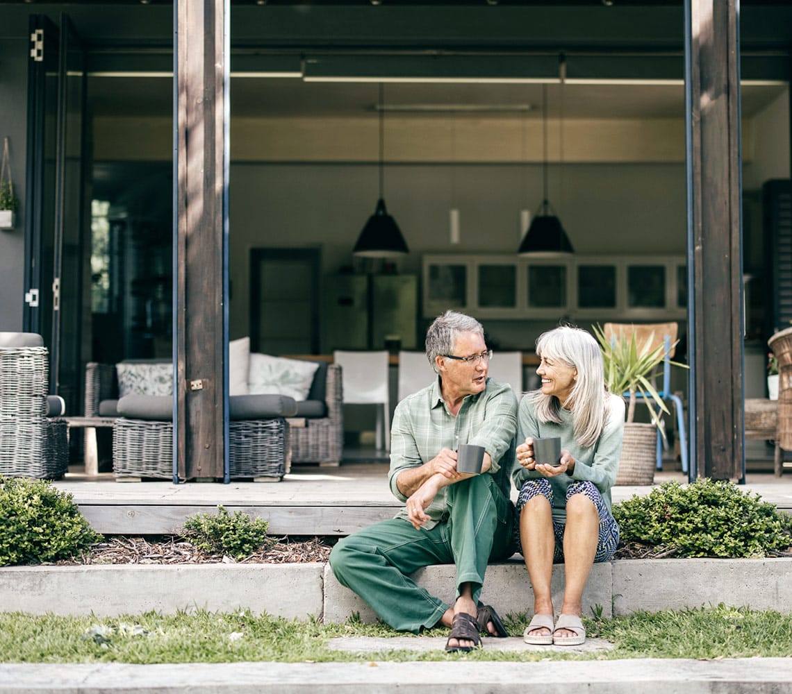An elderly couple sits on their home steps, holding coffee mugs and engaged in a conversation.